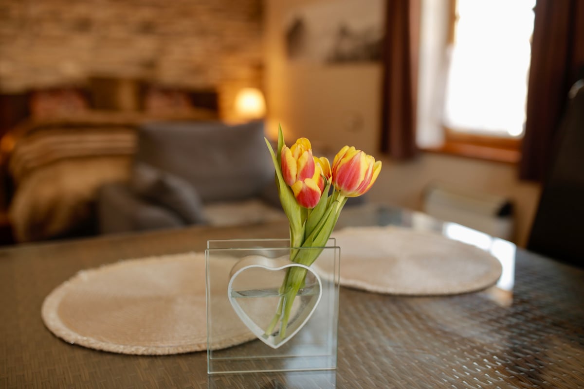 A small heart-shaped glass vase holds vibrant tulips on a round table. The background features a softly lit bedroom area with warm tones and rustic decor elements, creating a cozy ambiance. Natural light enhances the inviting space.