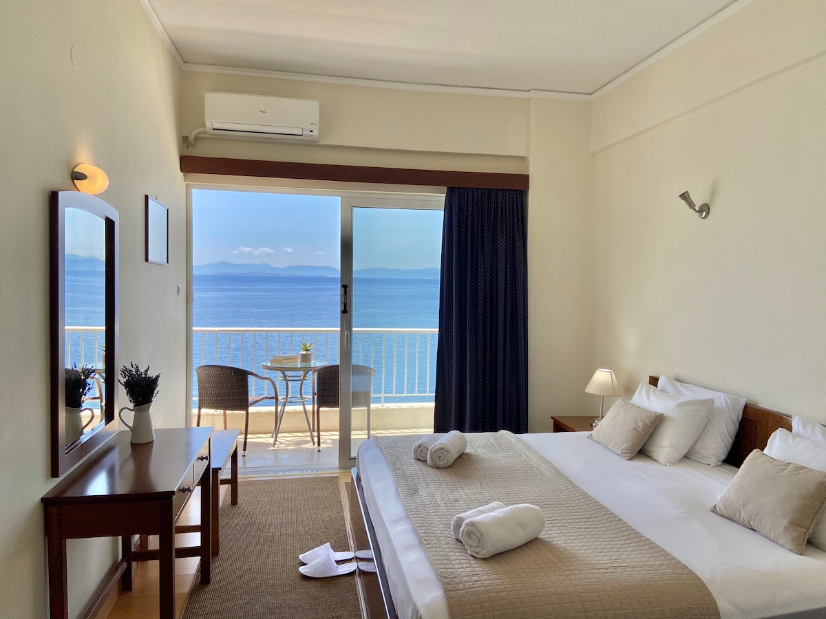 A bright room is shown with a spacious bed dressed in neutral linens. A balcony door provides a view of the sea and mountains in the distance. Two chairs and a table are placed on the balcony, enhancing the serene atmosphere of the oceanfront setting.