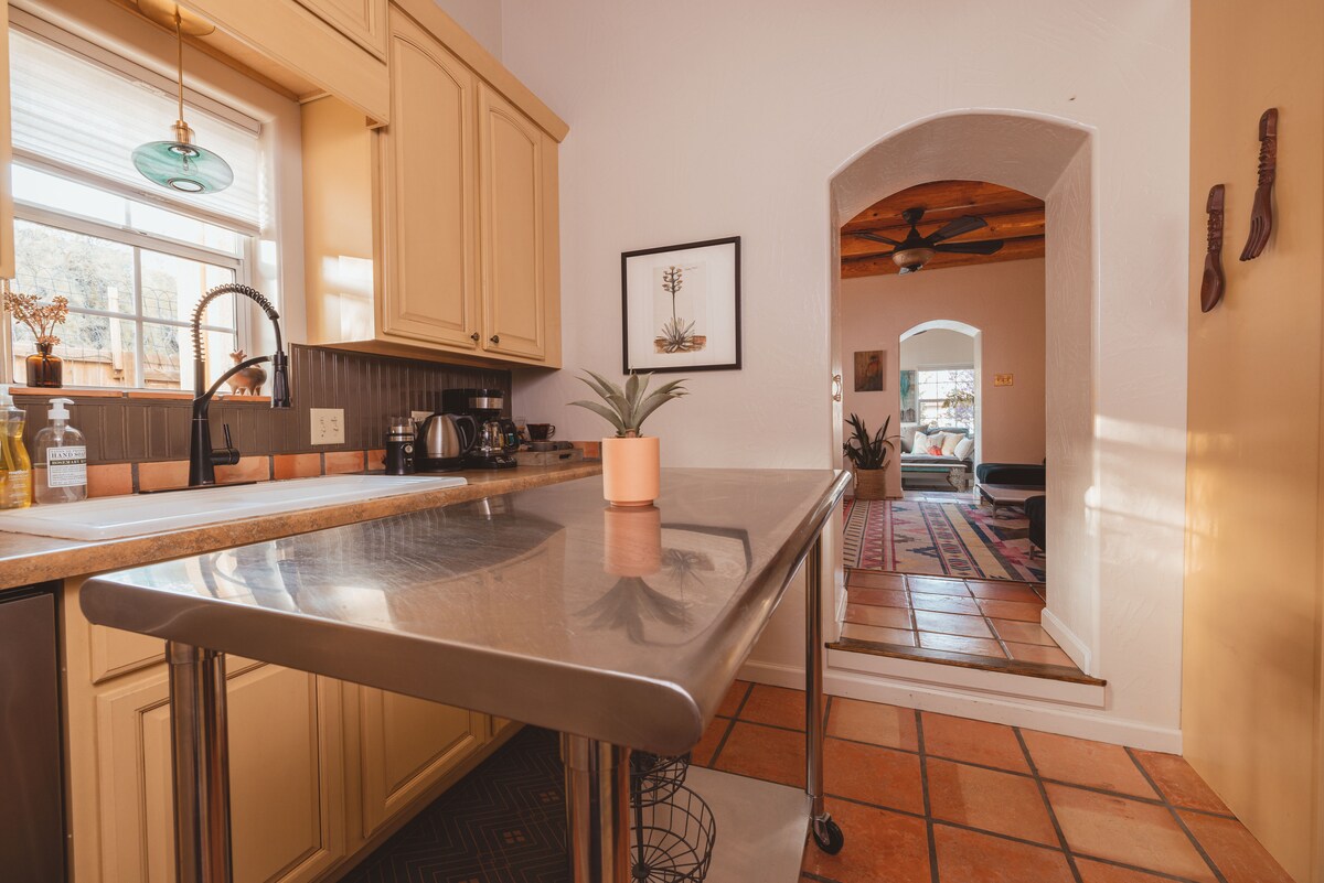 The kitchen features a stainless steel island on wheels, surrounded by custom cabinetry and modern appliances. An arched entryway leads to the living area, where warmth is provided by viga ceilings and a Kiva fireplace. Natural light enhances the inviting atmosphere.
