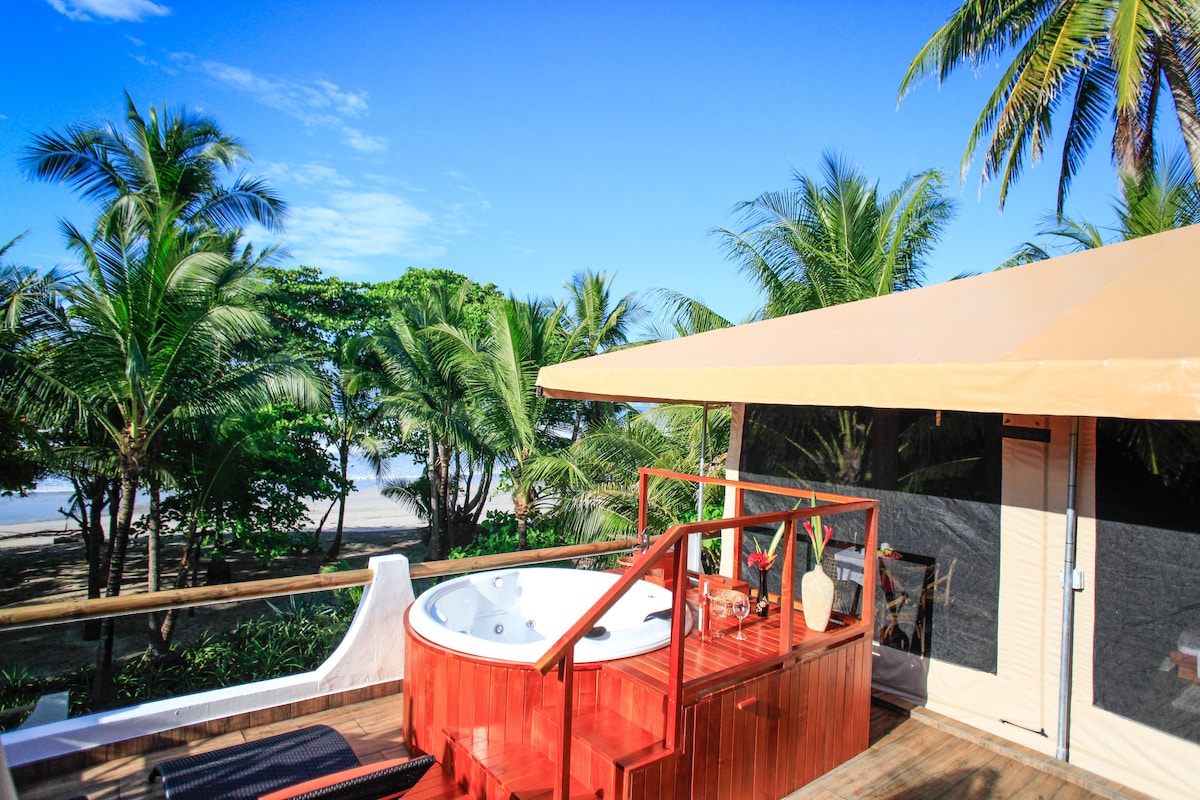 A private terrace features a wooden deck with a Jacuzzi for two, surrounded by lush palm trees. The space offers lounge chairs for relaxation and a shaded table, all set against a backdrop of clear blue skies and the distant ocean.