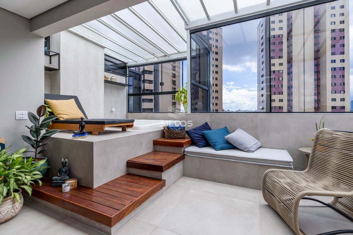 A covered leisure area features a relaxing jacuzzi and a comfortable seating space, accented by plush cushions. Wooden steps lead to a cozy nook surrounded by large windows, offering views of the surrounding buildings. Green plants add a touch of nature to the space.