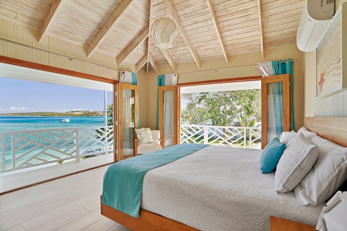 A spacious bedroom features a large bed with light-colored linens, accented by turquoise throw pillows. Large glass doors open to a stunning waterfront view, allowing natural light to flood the room. Light wooden beams create an airy atmosphere, complemented by turquoise curtains.