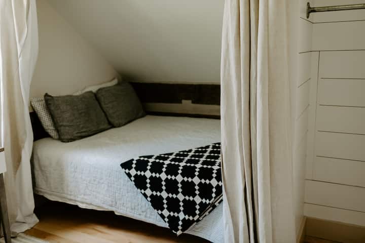 Cozy loft-style bedroom with a sloped ceiling, soft neutral bedding, and a bold navy-and-white patterned throw. A curtain offers privacy, making it a charming and intimate space for rest within the cabin’s warm, rustic atmosphere.
