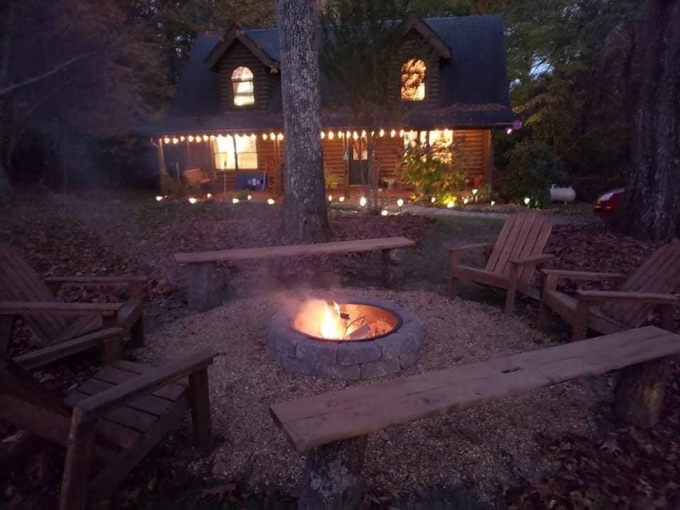 A fire pit surrounded by wooden benches is set in a gravel area, with gentle smoke rising towards the evening sky. The cozy cabin is illuminated by soft lights, showcasing large windows that invite warmth into the outdoor space.