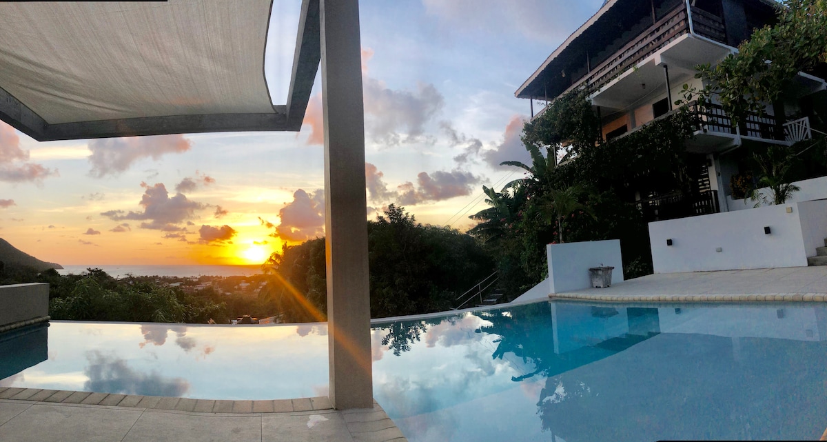 An infinity pool reflects the vibrant colors of a sunset over the ocean. The surrounding area is lush with tropical vegetation, while a multi-story building can be seen in the background, featuring balconies and outdoor spaces. The scene conveys a serene and inviting atmosphere.