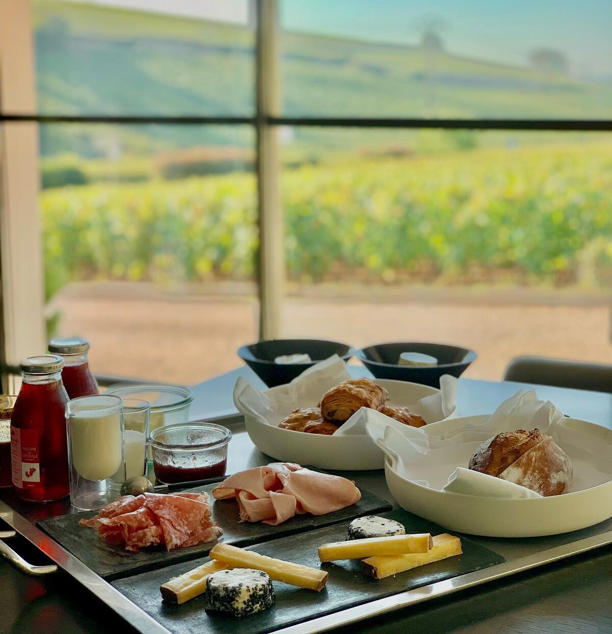 A breakfast spread is presented on a wooden tray, featuring assorted cheeses, sliced meats, pastries, and yogurt containers. In the background, large windows offer a view of lush vineyards, creating a serene dining atmosphere.