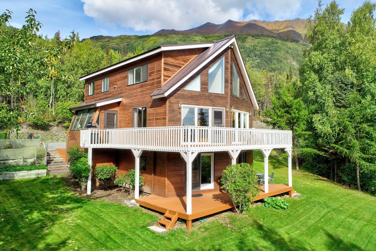 The two-story cabin features a rustic wooden exterior surrounded by lush greenery. A spacious wraparound balcony provides outdoor seating options and overlooks a grassy area. Large windows allow natural light to illuminate the interior, showcasing the inviting design against a backdrop of mountains.