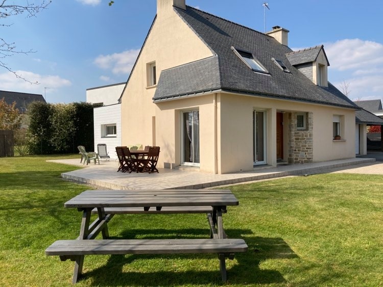 A spacious house is featured, complemented by a well-maintained garden with green grass. An outdoor dining area with wooden furniture is visible on the deck, while large windows invite natural light into the home. A patio area provides a welcoming space for relaxation.