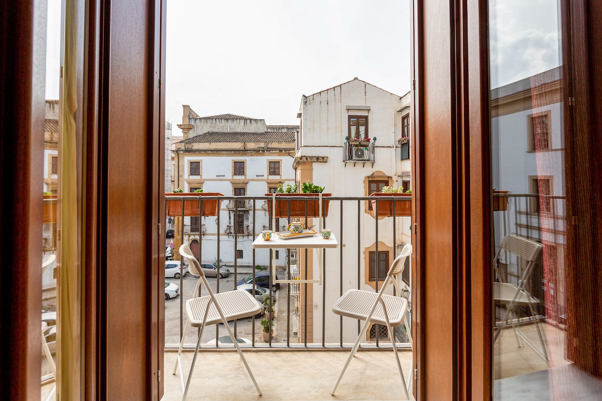 A small balcony is seen with two white folding chairs and a table set for two. Plants are placed on the balcony rail, providing a view of the historic buildings and streets below.