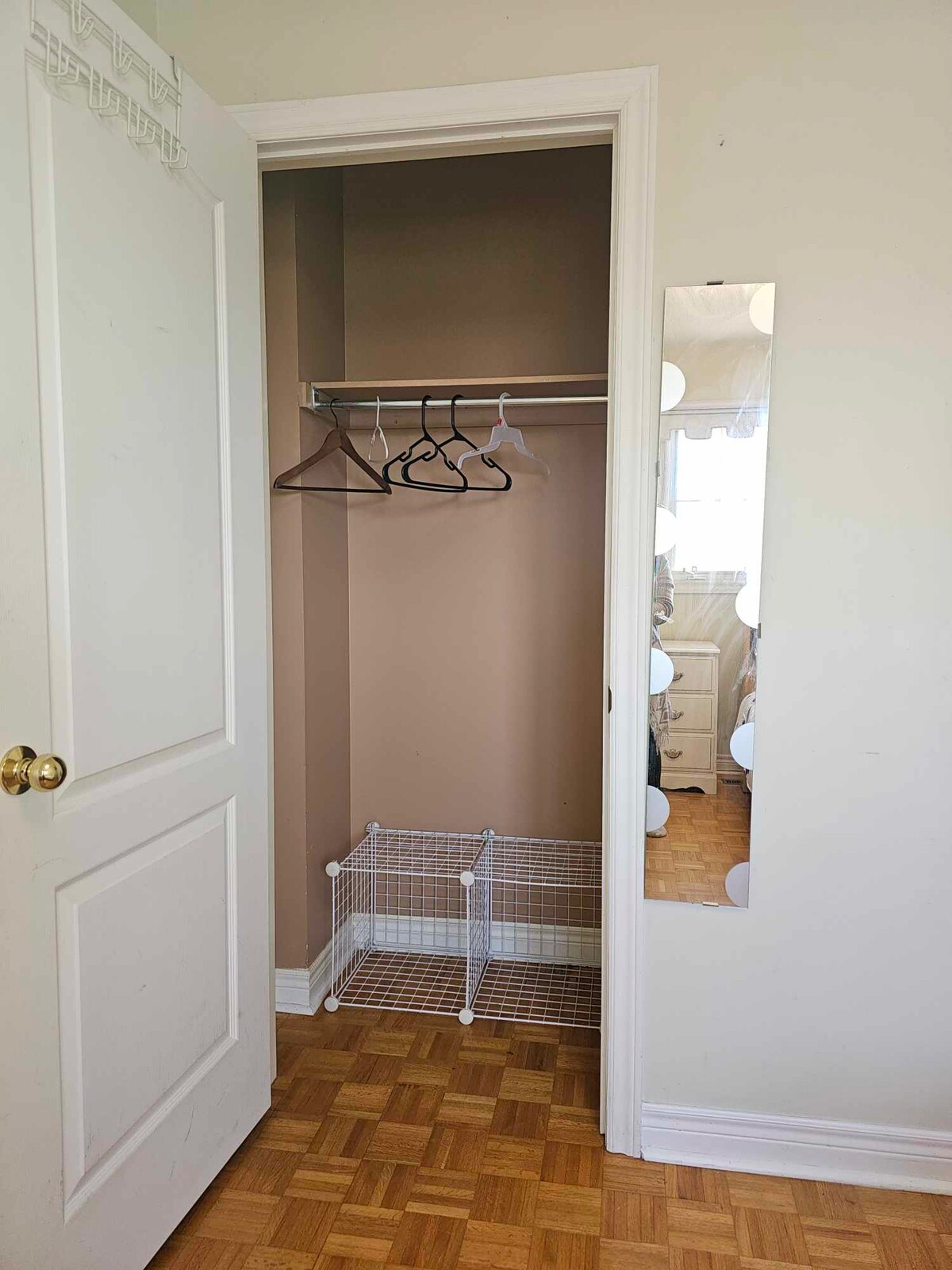 A small closet space is featured, highlighting a hanging rod with several hangers and a wire storage unit on the floor. A full-length mirror is visible on the adjacent wall, providing additional functionality. The walls are painted a soft neutral color, complementing the parquet flooring.