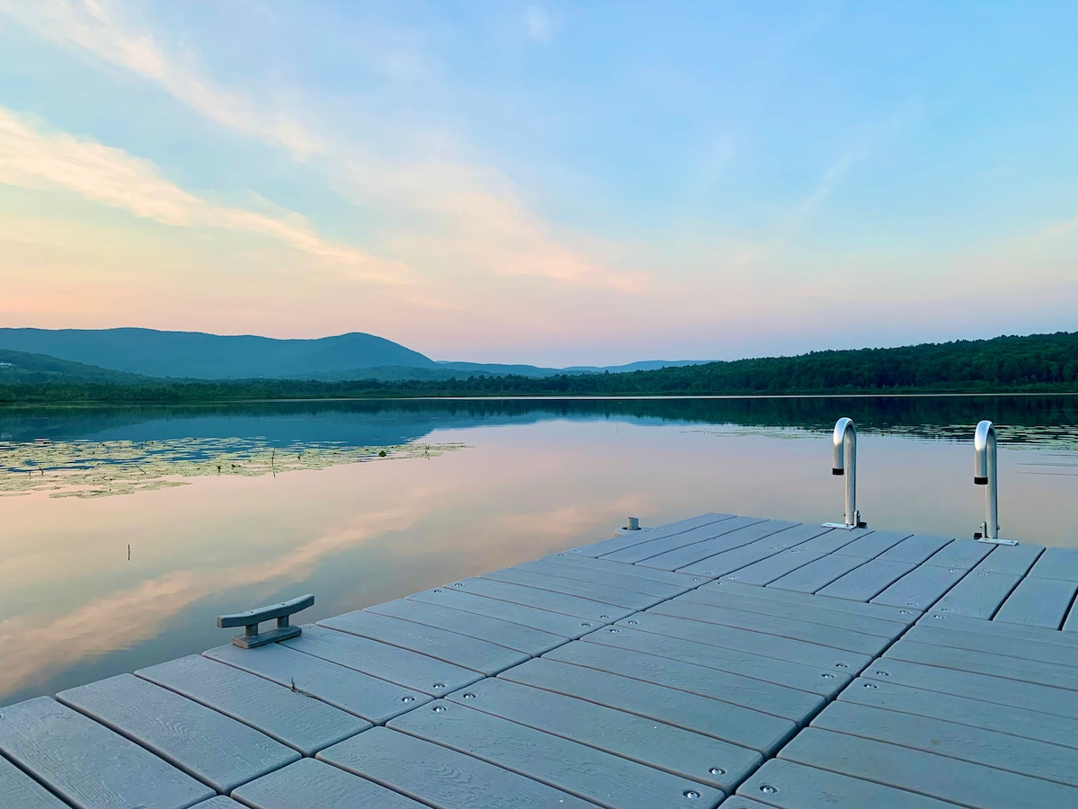 A wooden dock extends over calm waters, reflecting the pastel colors of the sky at dusk. Lush trees line the shore, while distant mountains are visible in the background, creating a serene and natural setting.