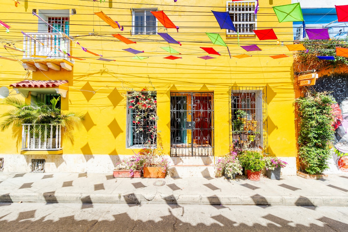 A vibrant yellow facade is adorned with colorful kites in shades of green, purple, and orange, casting playful shadows on the pavement. Potted plants and flowers line the entrance, and decorative detailing is visible on the window bars and balconies, enhancing the lively streetscape.