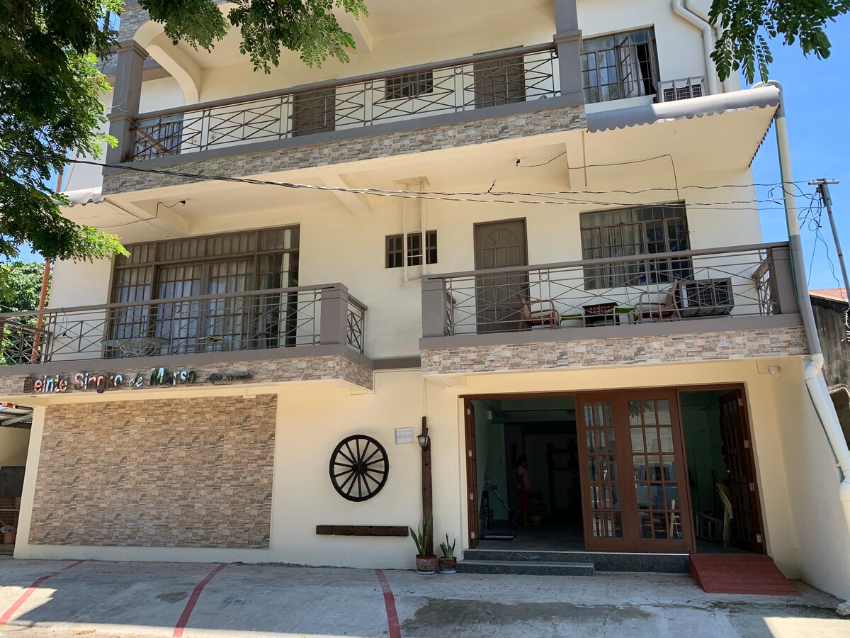 A three-story building features a mix of stone and stucco finishes. Large windows allow natural light to fill the interior spaces. A decorative wheel is mounted on the wall, and a few chairs and tables are positioned on the balcony, enhancing accessibility to the outdoors.