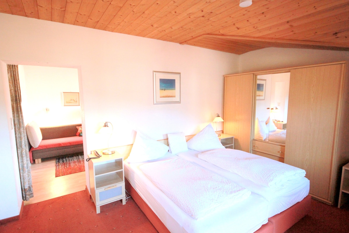 A well-lit bedroom is presented, featuring a spacious bed with white bedding and decorative pillows. A wooden ceiling adds warmth to the room. A mirror and a soft sofa are visible, complemented by two bedside lamps and a small side table.