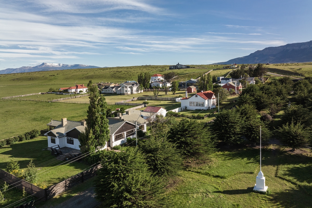 Aerial view of the scenic Puerto Bories neighborhood, featuring a cluster of houses with varied roof styles surrounded by green grass and trees. Majestic mountains rise in the background, with a clear blue sky above. The layout reflects a peaceful rural environment.