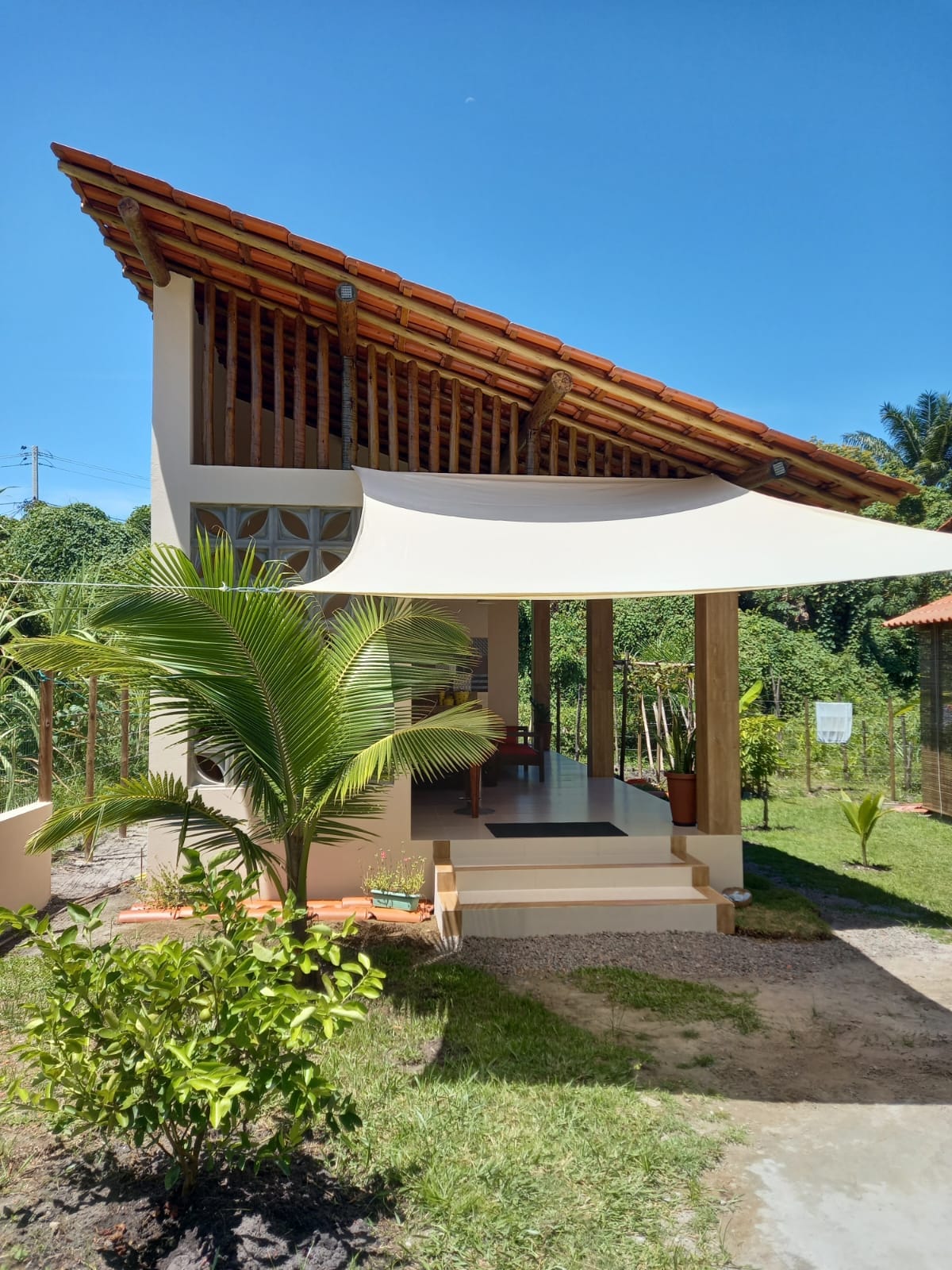 A bungalow with a sloped roof features a spacious front porch shaded by a large canopy. Lush greenery surrounding the property enhances the natural setting. The unique architectural elements, such as wooden beams and decorative blocks, add to the overall design while inviting the outdoors inside.