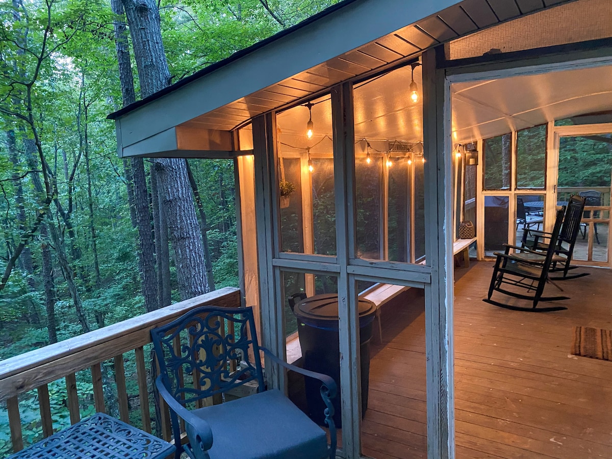 A screened porch surrounded by trees features multiple rocking chairs and warm string lights, creating a cozy outdoor space. Wooden flooring adds to the inviting atmosphere, while nature's greenery is visible through the mesh screens, providing a serene view.