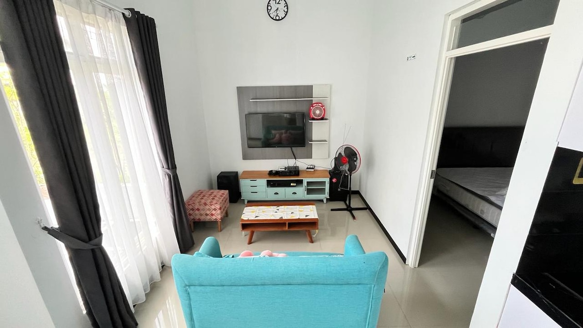 A living area is presented with a pastel blue sofa and a low wooden coffee table. A wall-mounted smart TV is visible, accompanied by a fan and a small red pouf. Sheer curtains frame the window, allowing natural light to fill the space.