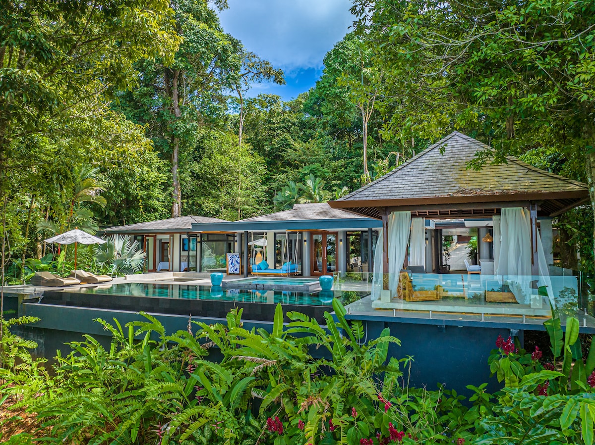 The villa is nestled amidst lush tropical foliage, featuring an outdoor area with a private infinity pool and elegant seating. Large glass doors open to an airy living space, blending seamlessly with nature. Sun loungers and an umbrella provide options for relaxation.