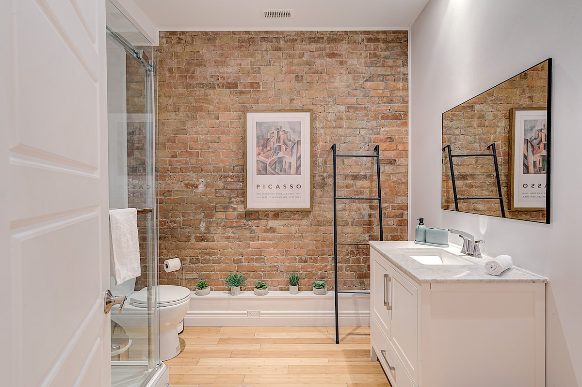 A modern bathroom features exposed brick walls and a large mirror above a sleek vanity. A glass shower is visible, alongside neatly arranged plants on the countertop. Towels are positioned neatly, enhancing the clean and organized look of the space.