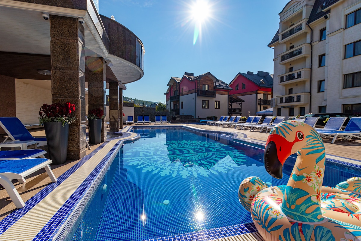 A large outdoor swimming pool is featured, surrounded by sun loungers and vibrant floral planters. A colorful float shaped like a flamingo rests on the water's surface. Sunlight reflects off the pool, creating a bright and inviting atmosphere in the swimming area.