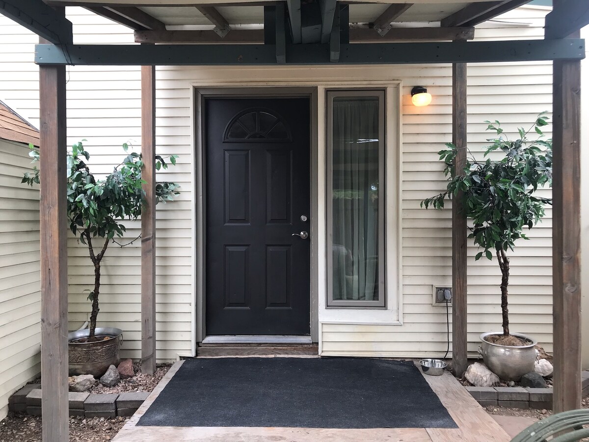The entrance features a black front door under a covered porch, framed by two potted plants. A welcome mat is positioned in front, and a light fixture provides illumination. The exterior is characterized by light-colored siding and a simple, tidy layout.