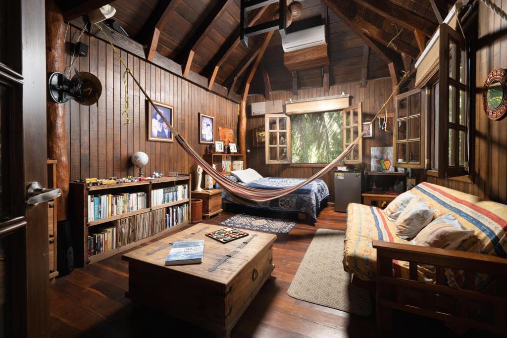 A rustic cabin interior features wooden walls and a high ceiling. A spacious bed is positioned beside a hammock, while a small sofa extends across the room. A bookshelf and coffee table add character, and windows allow natural light to illuminate the space.