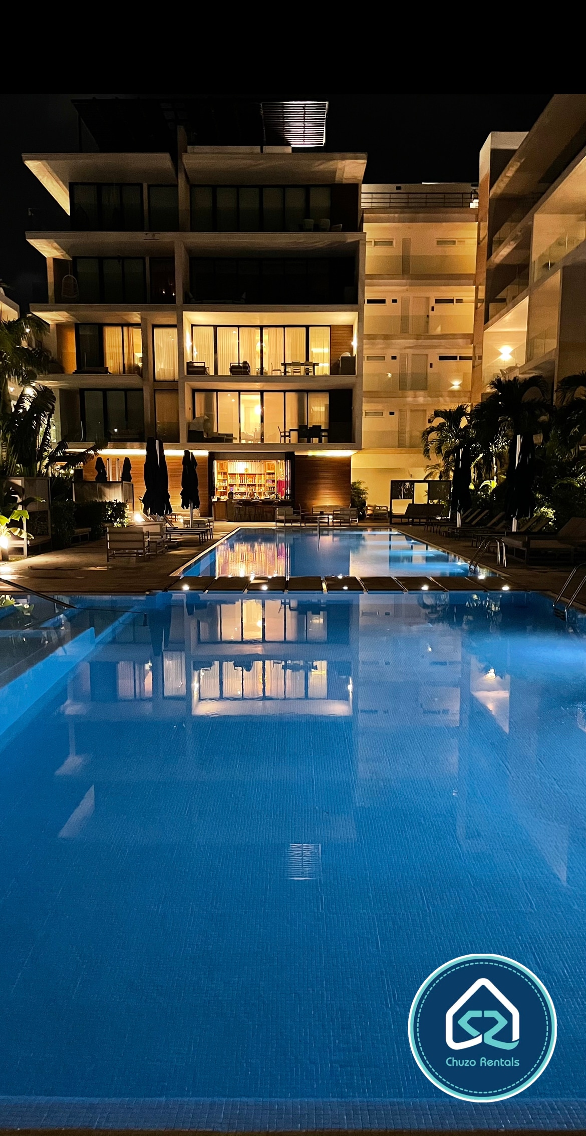 A modern heated pool reflects the surrounding lighting, creating a serene atmosphere. Lounge chairs line the pool’s edge, and a multi-story building with large glass windows is visible in the background, enhancing the contemporary view.