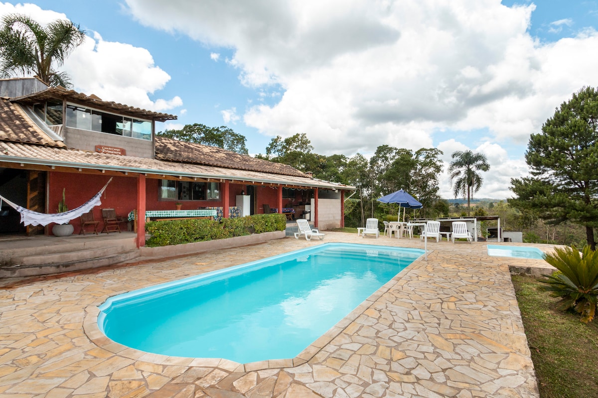 A clear blue pool is surrounded by a stone patio, with lounge chairs and a parasol positioned nearby. The rustic house, featuring open spaces and large windows, is set against a backdrop of lush greenery and a partly cloudy sky.