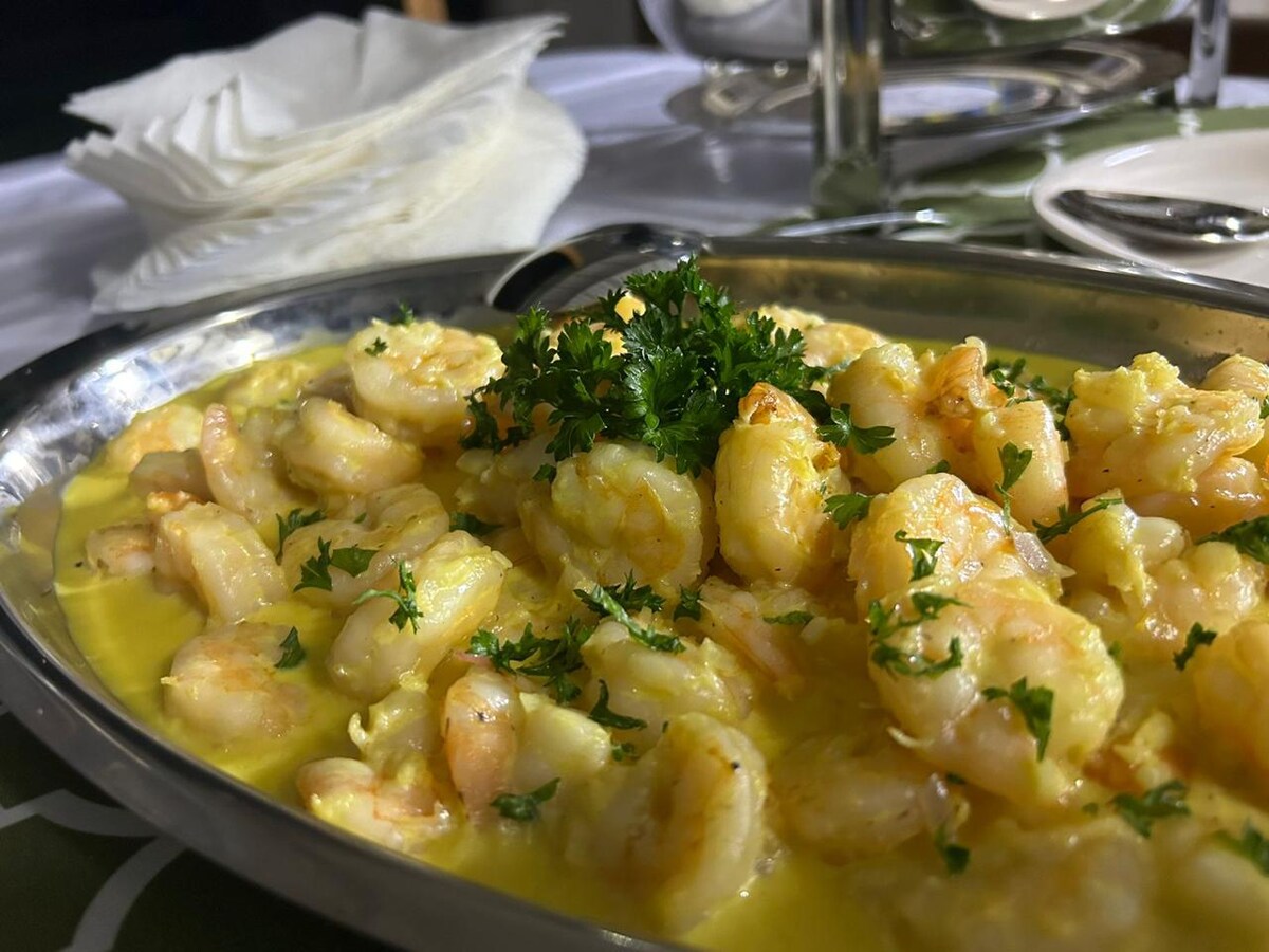 A stainless steel platter is filled with lightly sautéed shrimp, garnished with fresh green parsley. A creamy yellow sauce pools around the shrimp, adding color and richness to the dish. In the background, white plates and napkins are arranged on a table set for dining.