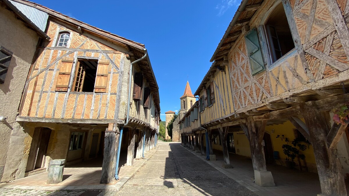 An historic village street showcases half-timbered houses with wooden beams and ornate shutters. The buildings are elevated on stilts, framing a central pathway that leads towards a church with a pointed steeple visible in the background under a clear blue sky.