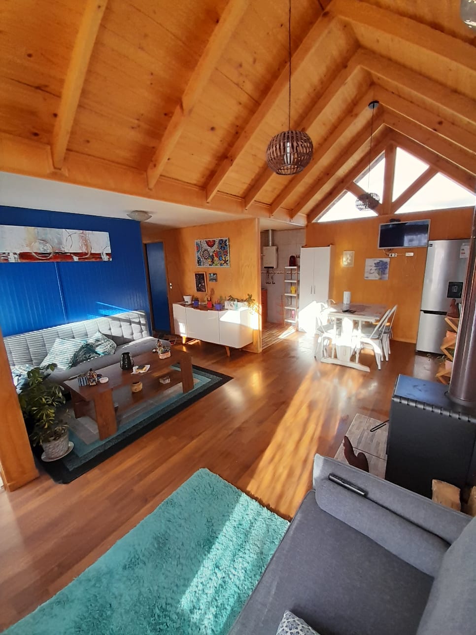 The interior space features high wooden ceilings with beams and skylights that allow natural light to illuminate the area. A cozy living area with stylish seating is complemented by a dining space and a well-equipped kitchen in the background.