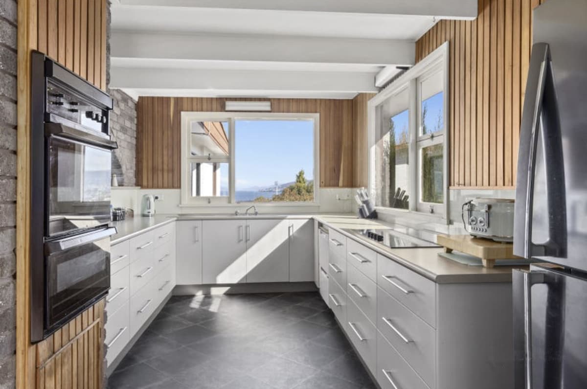 A modern kitchen with sleek white cabinetry and stainless steel appliances is displayed. Natural light filters through large windows, highlighting the wooden paneling on the walls. The gray tile flooring adds a contemporary touch to the overall design.