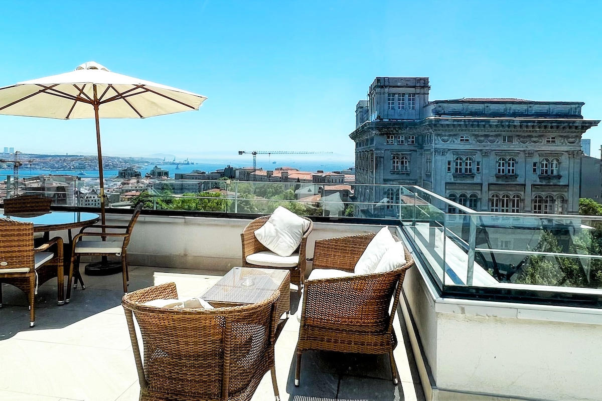 A spacious terrace features a seating area with wicker furniture, including chairs and a coffee table. An umbrella provides shade while panoramic views of the Bosphorus and surrounding architecture are showcased, highlighting the outdoor space's connection to the vibrant city.