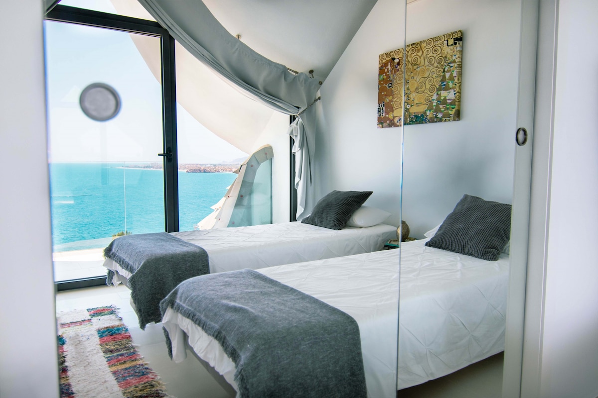 Dormitorio con camas individuales, ropa de cama blanca, cojines y mantas decorativas, toallas, alfombra con diseño, cuadro en pared y armario con espejos. Ventanal acristalado con cortinas que conecta con terraza de cristal y ofrece vistas al mar.