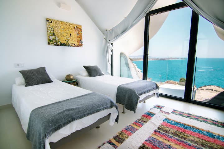 Bedroom with single beds, white linens, cushions and decorative blankets, towels, carpet with design, picture on the wall and wardrobe with mirrors. Glazed window with curtains that connects to a glass terrace and offers sea views.