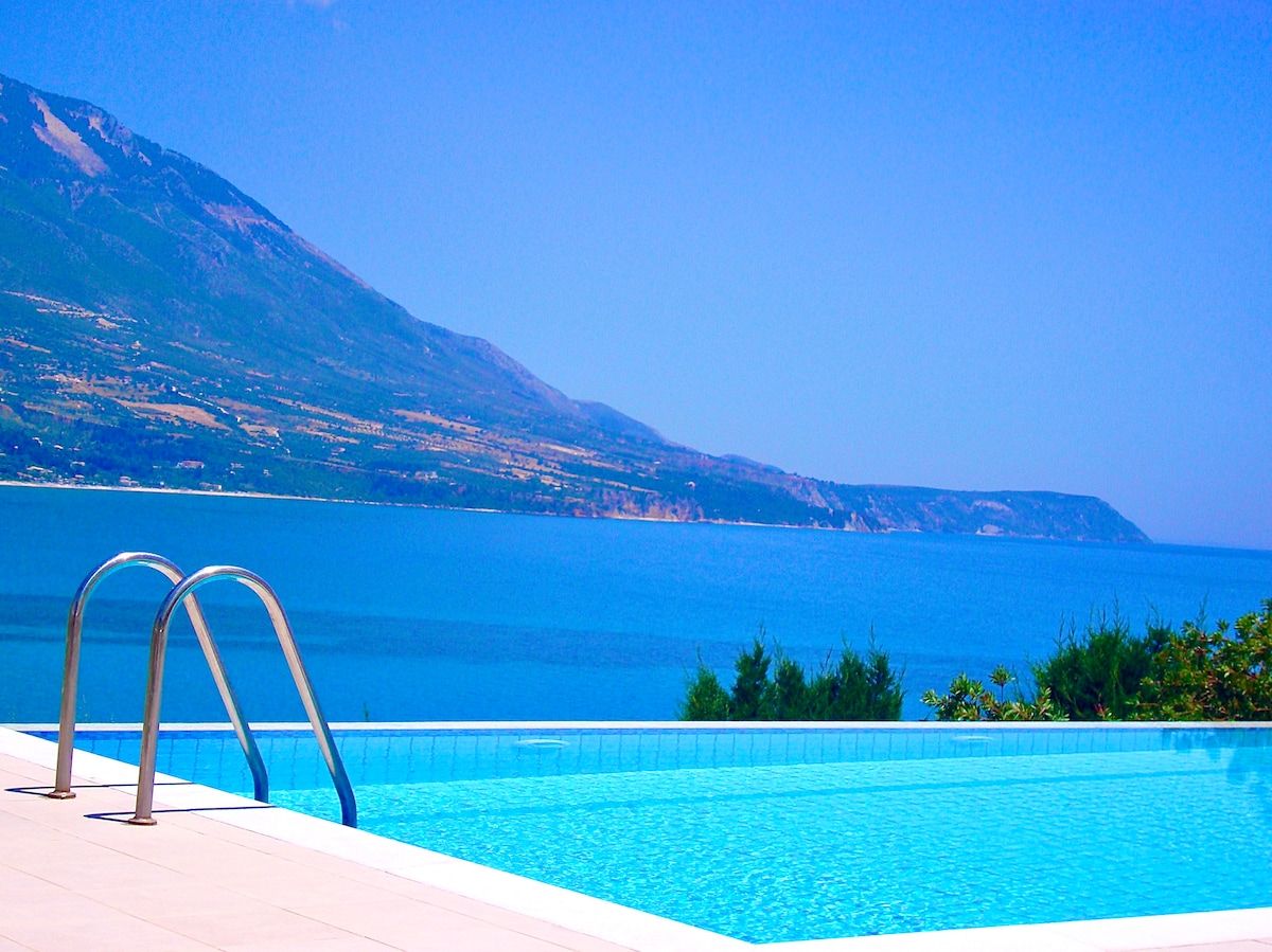 An infinity swimming pool extends towards the horizon, reflecting the clear blue sky and the vast sea. Lush greenery borders the pool area, and mountains can be seen in the distance, adding to the serene coastal ambiance.
