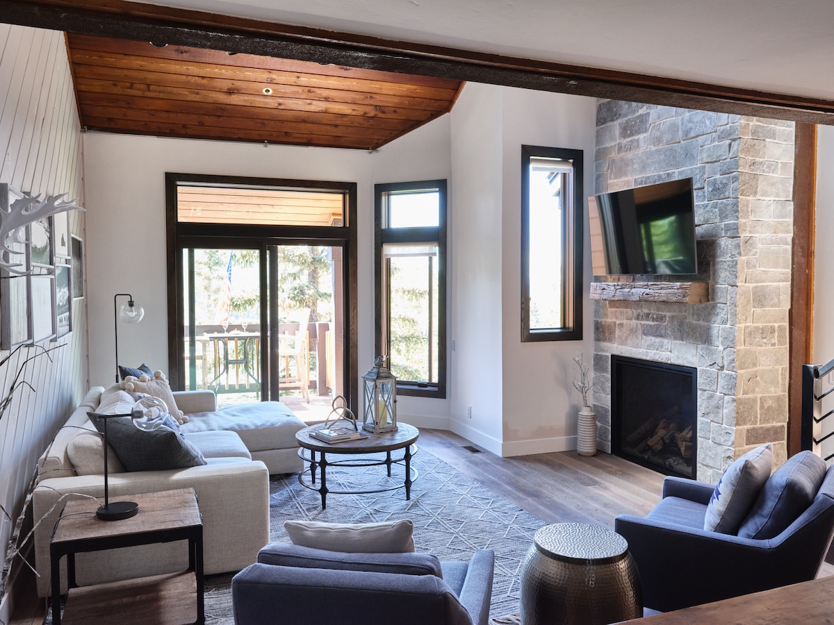 The living area is adorned with a combination of light and dark furnishings. A stone fireplace anchors the space, and large windows frame views of the outdoors. A cozy sofa and armchairs are arranged around a central coffee table, creating an inviting atmosphere.