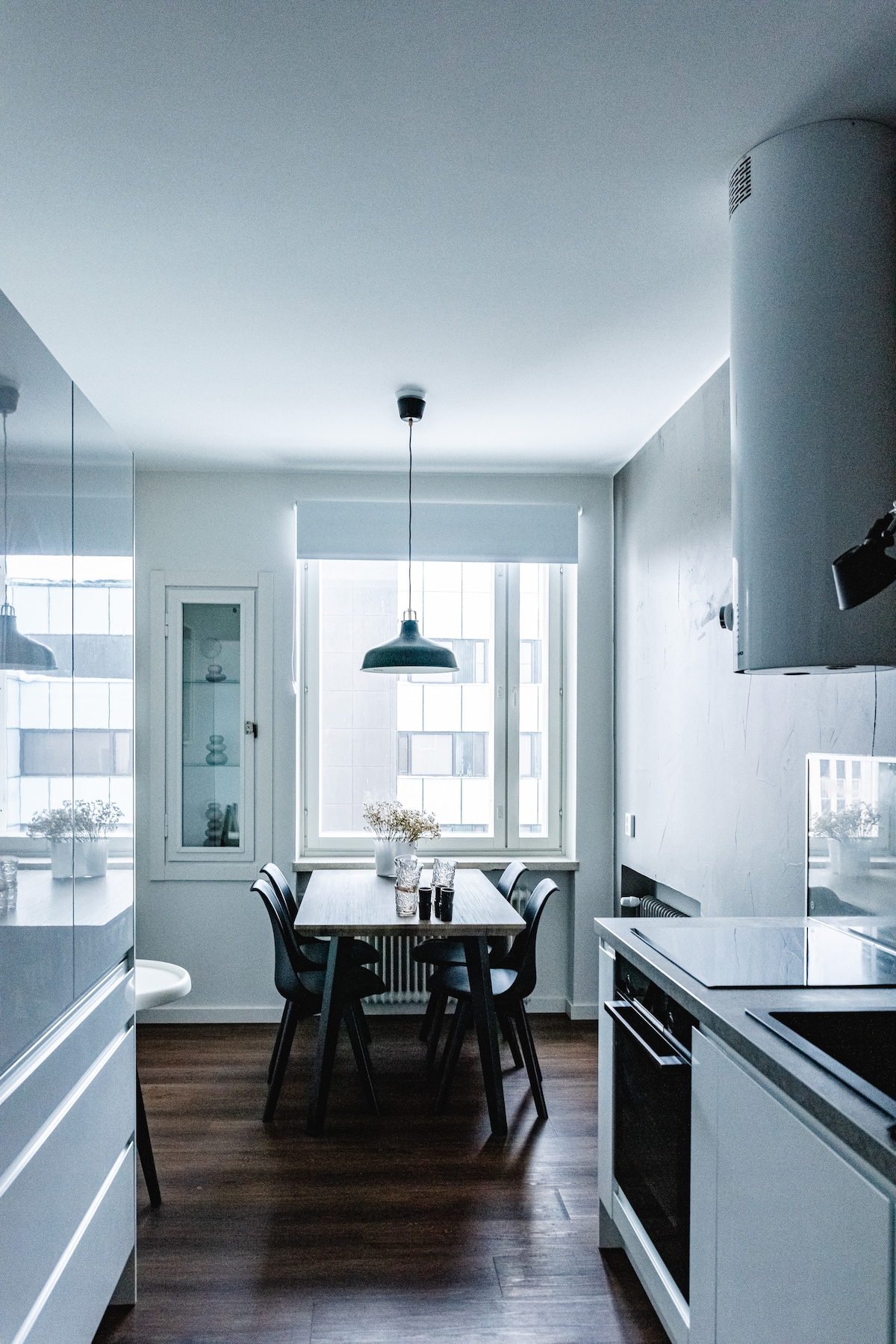 A bright kitchen area features a wooden dining table surrounded by black chairs, illuminated by a pendant light. Tall windows allow natural light to flood the space, highlighting the modern cabinetry and countertop appliances. Neutral-colored walls contribute to a clean and inviting atmosphere.