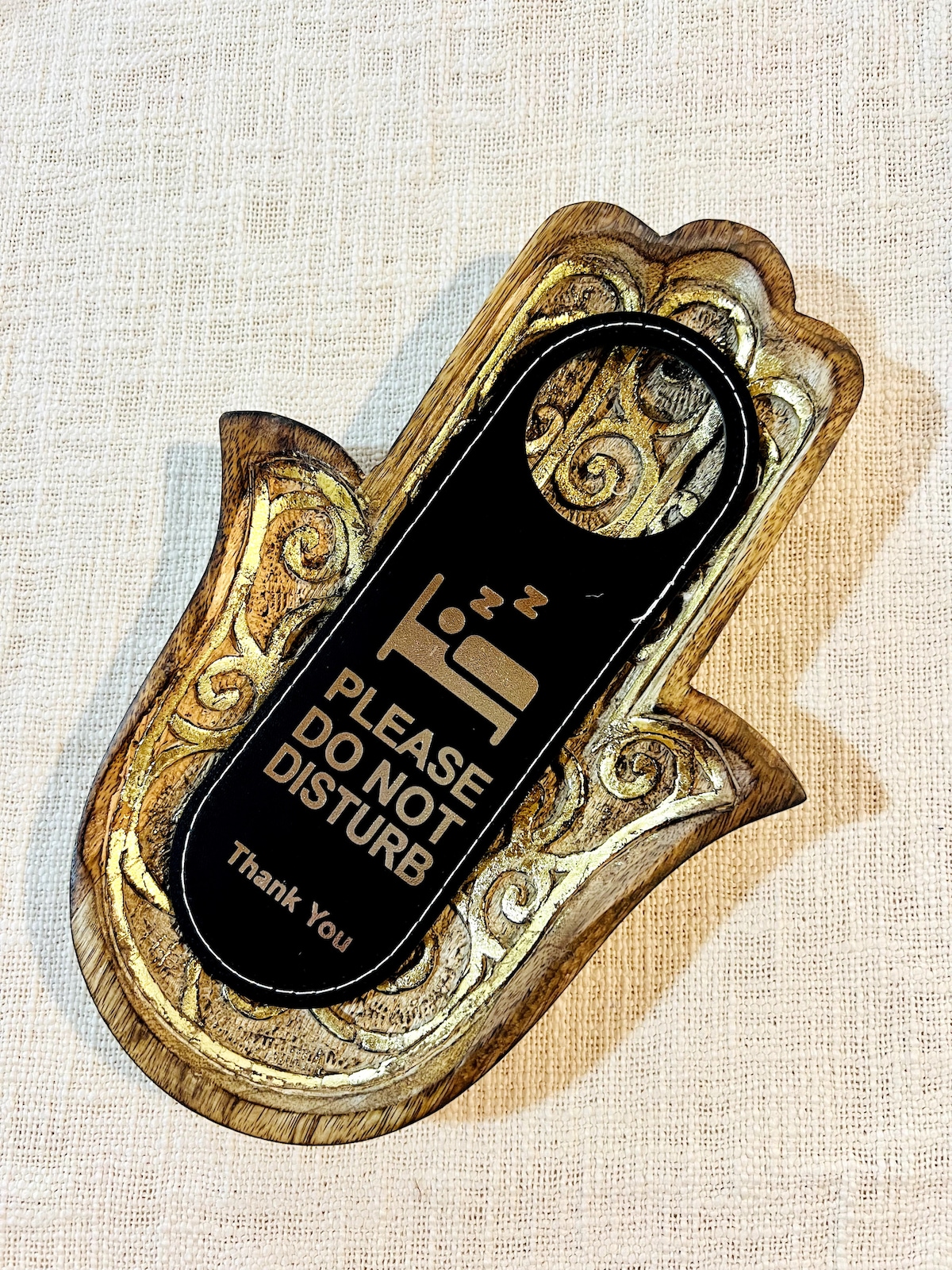 A decorative door hanger is displayed, shaped like a hand. The wooden piece features ornate details, with a black insert stating 'PLEASE DO NOT DISTURB' and an image of a sleeping person. The lower part includes the words 'Thank You' in a smaller font.