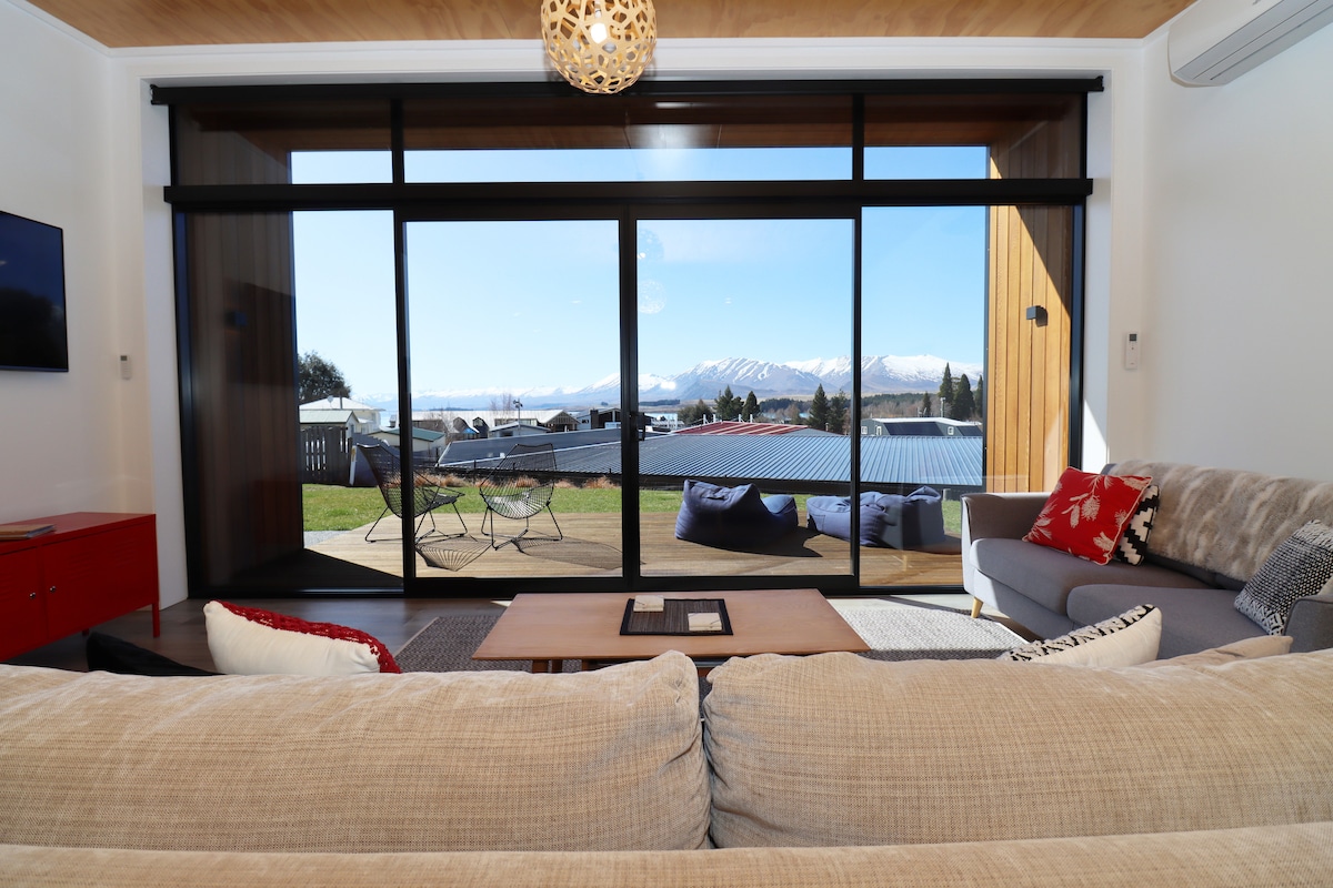 A spacious living area is showcased, featuring large glass doors that open to reveal expansive views of the mountains. Comfortable seating, including a grey sofa and a patterned chair, invites relaxation. Natural light illuminates the room, highlighting the warm wooden ceiling and modern decor.