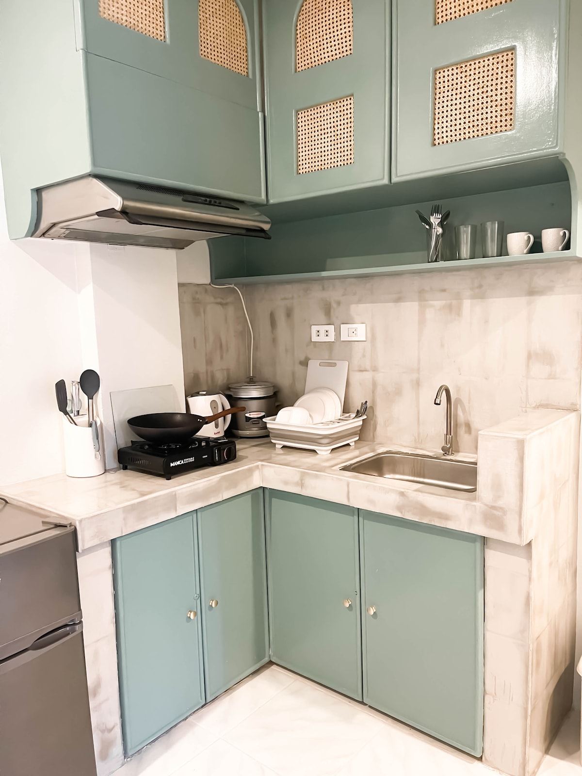 A well-equipped kitchen features mint green cabinetry and a marble countertop. A stove, sink, and drying rack are visible, alongside a stainless steel refrigerator. Essential kitchen utensils and dishware are neatly arranged, providing a functional space for cooking and meal preparation.