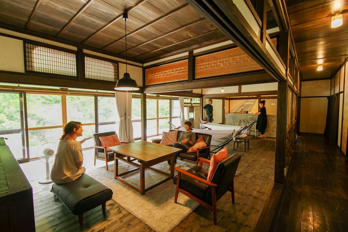 The interior space features a traditional design with wooden beams and a cozy seating area. A central table is surrounded by comfortable chairs. Large windows provide ample natural light and views of the greenery outside, creating an inviting environment for relaxation.