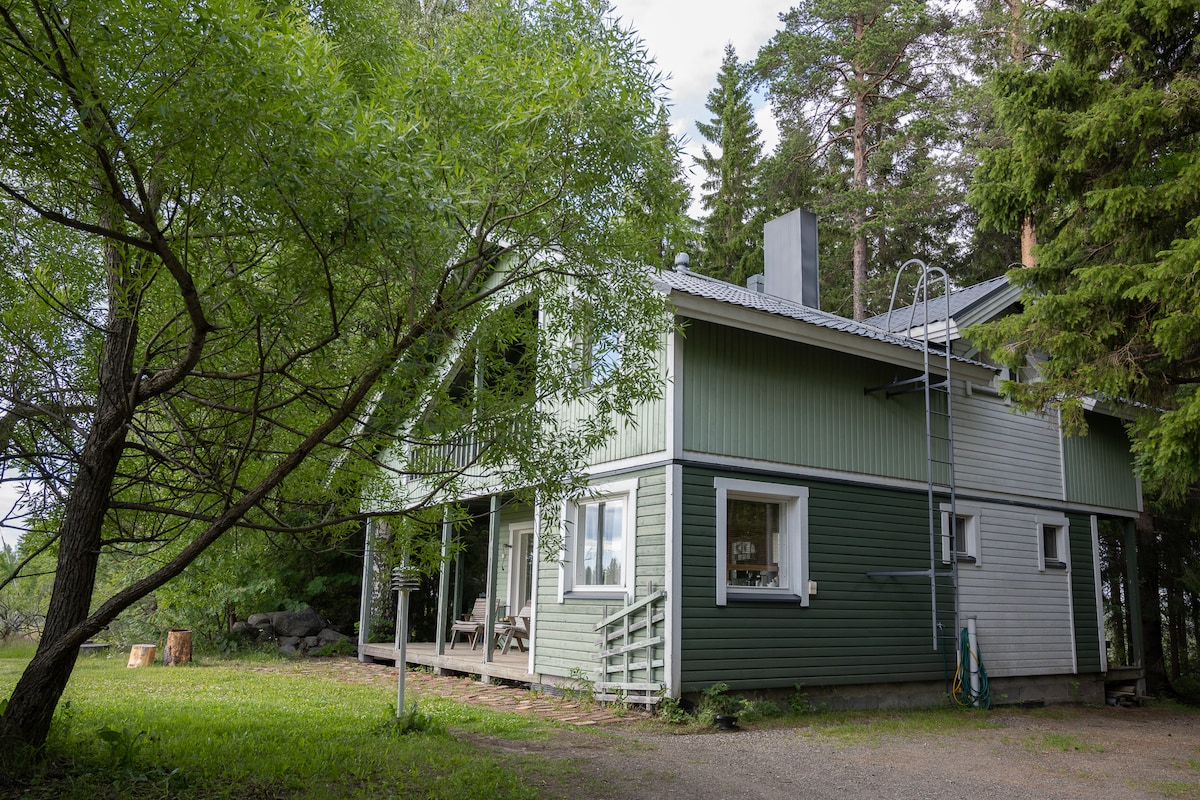 The exterior of the villa is shown, featuring a combination of green and white cladding. Surrounding trees offer a sense of privacy. A small porch is visible at the front, providing outdoor seating. A ladder leans against the building, indicating access to the roof area.