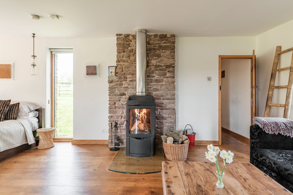 A warm wood burner is positioned on a stone feature wall, providing a cozy focal point in the spacious interior. A mix of seating options and natural light from a nearby window create an inviting atmosphere. The wooden flooring complements the rustic charm of the space.