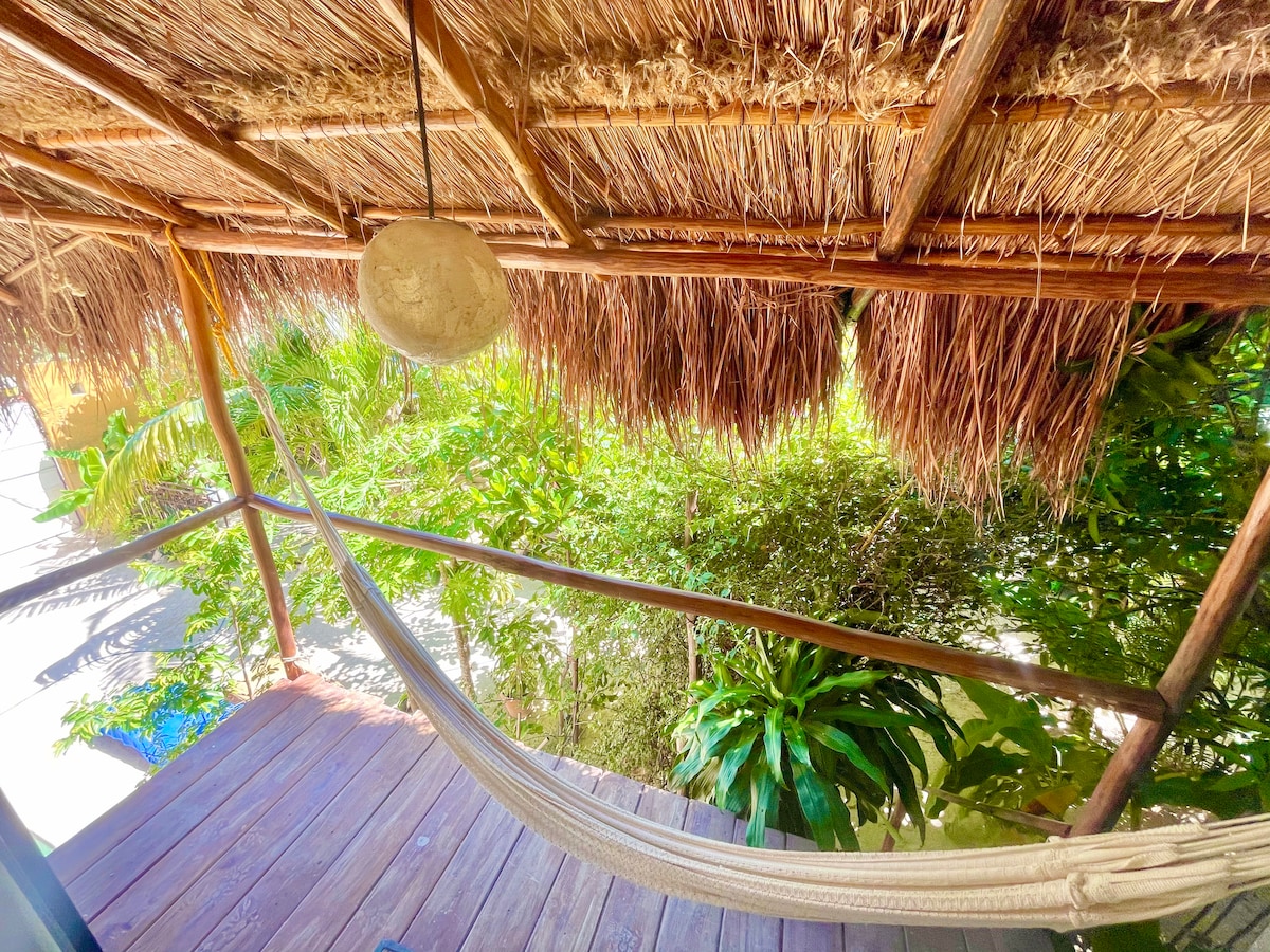 A private deck features a hammock suspended beneath a thatched roof. Lush greenery surrounds the area, creating a serene atmosphere. Wooden flooring enhances the natural aesthetic, while the sun casts warm light on the foliage, inviting moments of relaxation.