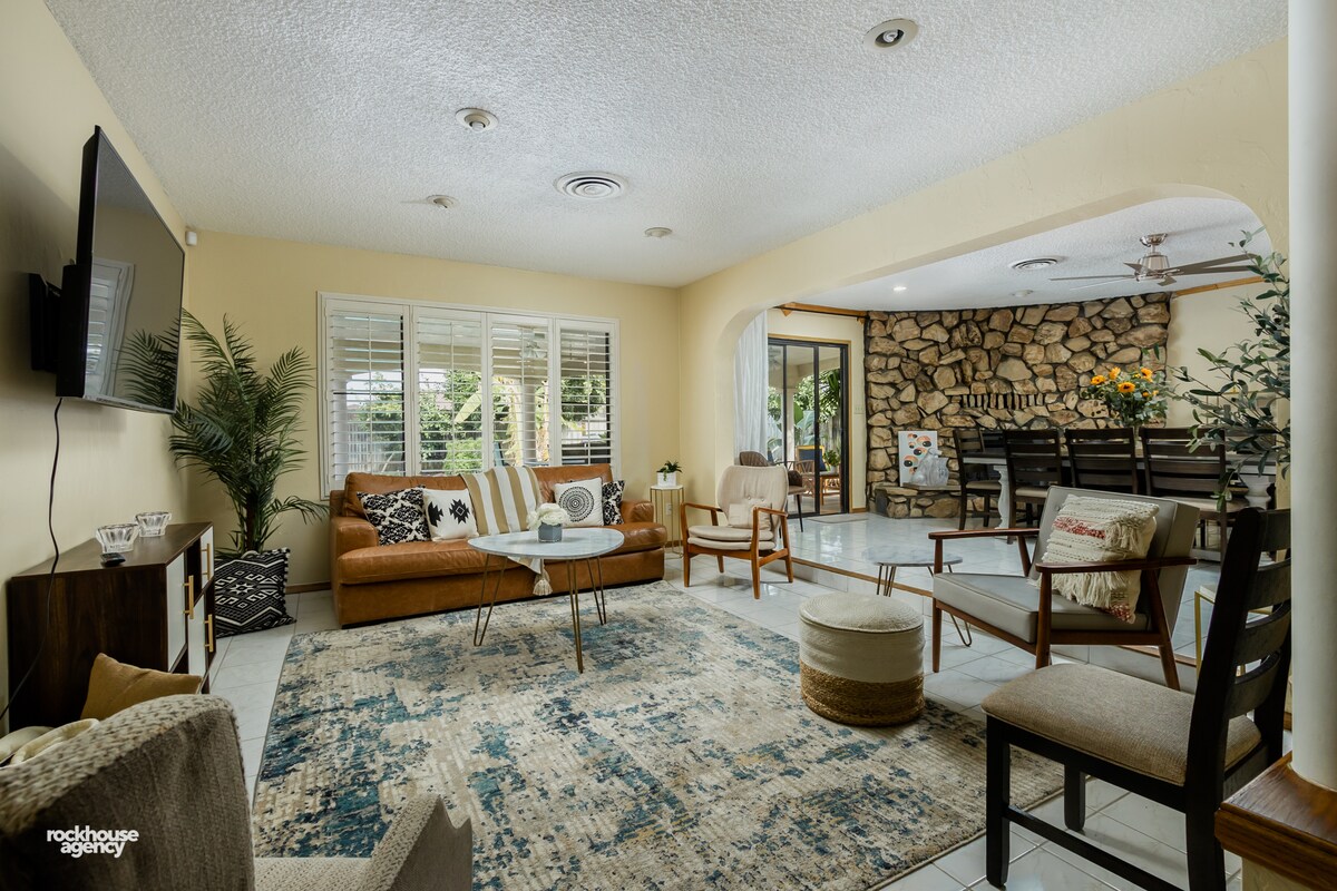 A cozy living area features a brown leather sofa and armchairs arranged around a central coffee table. A decorative rug lies beneath, and large windows allow natural light to fill the space. A stone accent wall adds character, complementing the warm tones throughout.