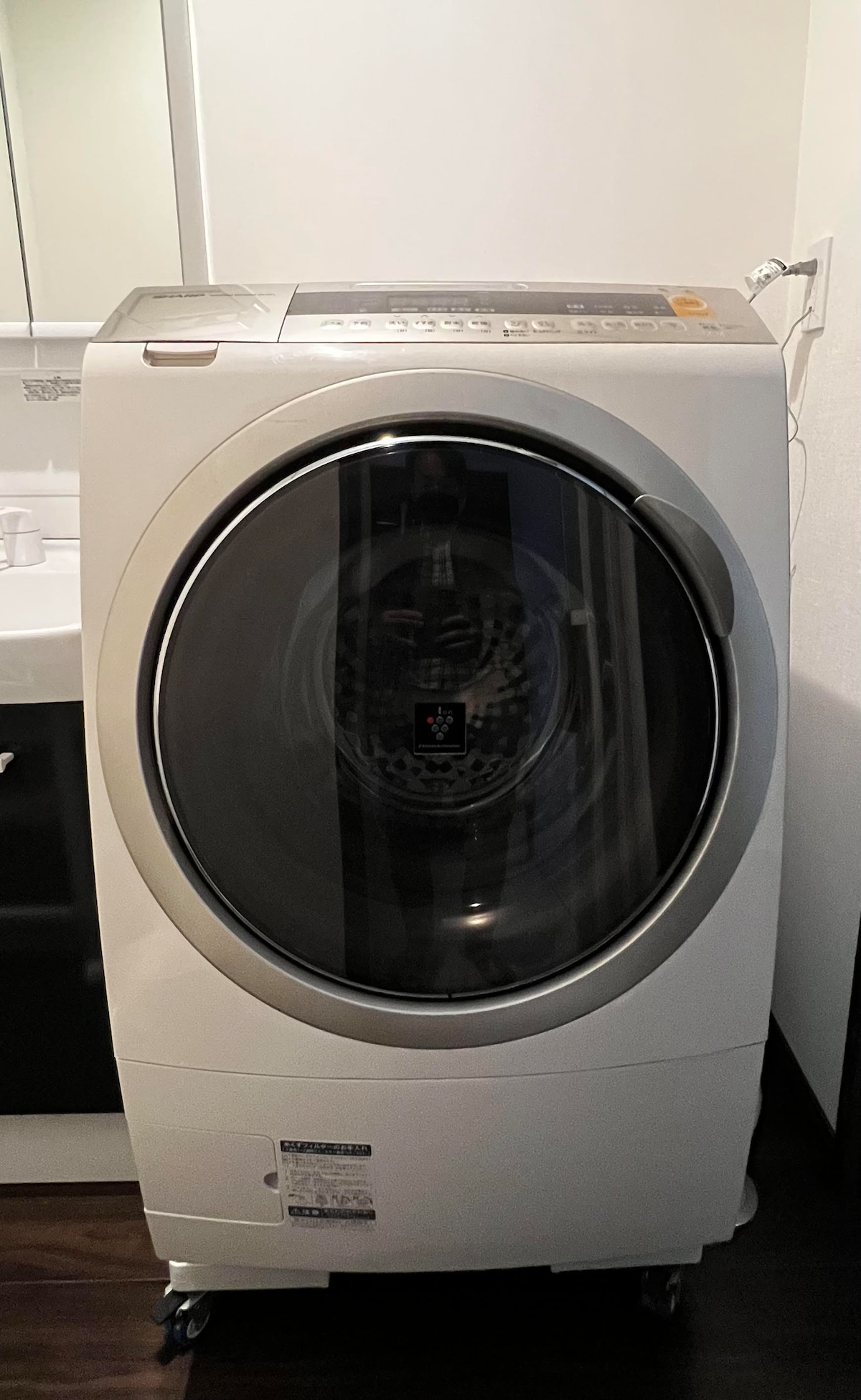 A washing machine with a large transparent door is positioned against a neutral wall. The machine is equipped with various controls on its top panel, allowing for easy selection of washing options. A well-lit space is provided nearby for utility.