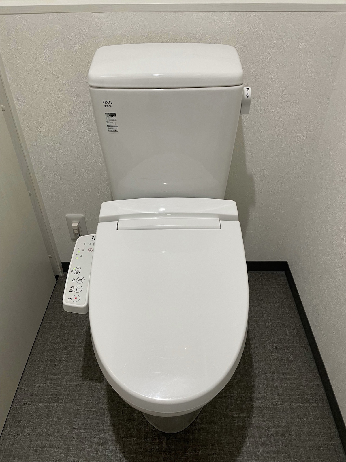 A modern toilet is positioned against a plain wall, featuring a sleek design. Controls for various functions are located on the side. The floor is covered in neutral-colored tiles, contributing to a clean and tidy appearance.