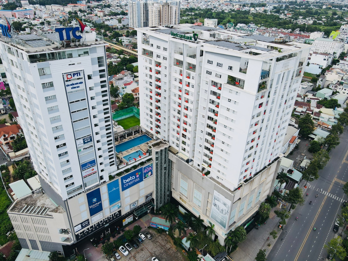 An aerial view captures The Pegasus Plaza with its two distinct towers, showcasing a mix of residential and commercial spaces. The surrounding area features streets lined with greenery and a glimpse of the rooftop swimming pool. The cityscape extends into the background with various buildings.